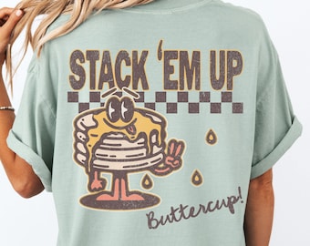 May include: A light green t-shirt with a graphic of a cartoon pancake stack with the text "Stack 'Em Up Buttercup!" printed on the back.