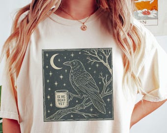 May include: A white t-shirt with a black and white illustration of a raven perched on a branch with a mug that says "Is he dead yet?" The background is a dark blue with stars and a crescent moon.