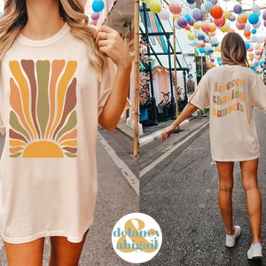 May include: Cream-colored t-shirt featuring a retro-style sunburst design in shades of orange, brown, and green. The back of the shirt displays the text "forever chasing sunsets" in a matching color palette. A casual, comfortable tee.
