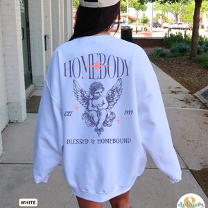 May include: White sweatshirt with a graphic design on the back. The design includes the word "HOMEBODY" above an angel illustration with wings, the text "EST 1999", and the words "BLESSED & HOMEBOUND".