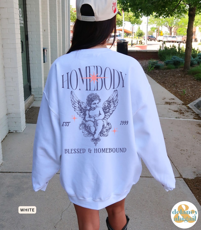May include: White sweatshirt with a graphic design on the back. The design includes the word "HOMEBODY" above an angel illustration with wings, the text "EST 1999", and the words "BLESSED & HOMEBOUND".