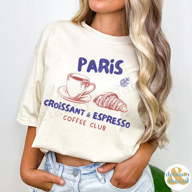 May include: A beige t-shirt with a graphic design that says "Paris Croissant & Espresso Coffee Club". The graphic features a cup of coffee, a croissant, and a leaf.