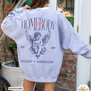 May include: Sports grey sweatshirt with the word "HOMEBODY" above an angel graphic. Below the angel are the words "BLESSED & HOMEBOUND", with "EST" and "1999" on either side. The sweatshirt has a white collar.
