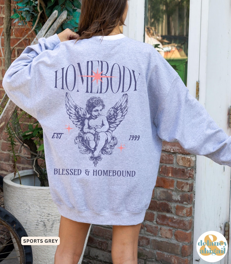 May include: Sports grey sweatshirt with the word "HOMEBODY" above an angel graphic. Below the angel are the words "BLESSED & HOMEBOUND", with "EST" and "1999" on either side. The sweatshirt has a white collar.