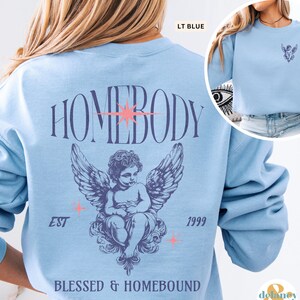 May include: Light blue sweatshirt with the word "HOMEBODY" arched across the back. Below the text is a detailed illustration of a winged cherub. The text "EST 1999" and "BLESSED & HOMEBOUND" are also printed on the sweatshirt.
