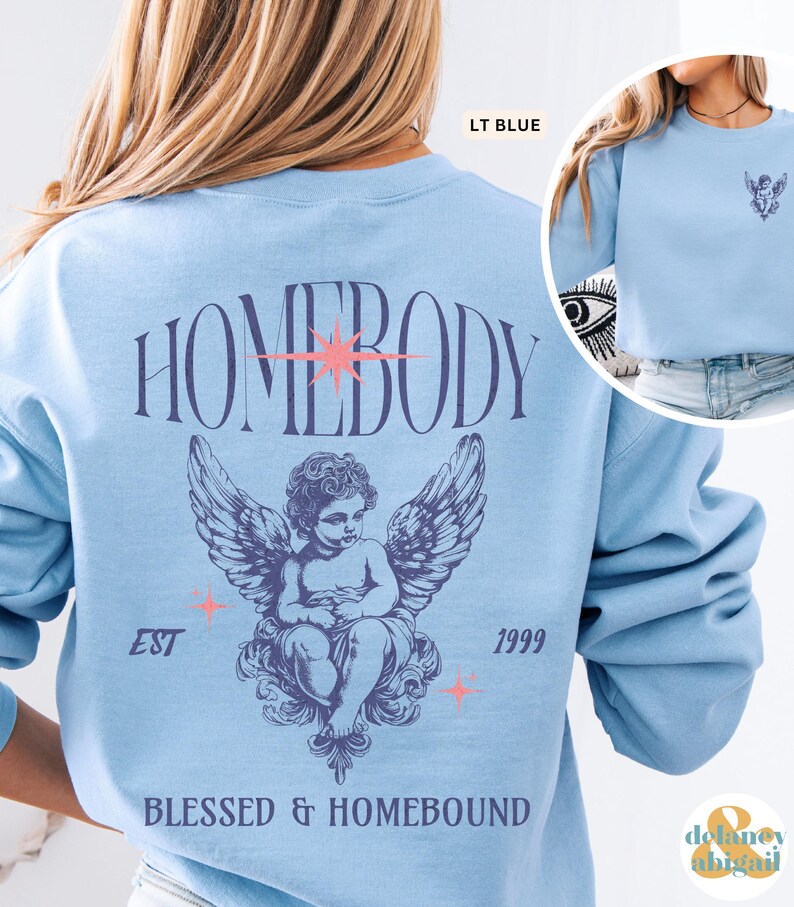 May include: Light blue sweatshirt with the word "HOMEBODY" arched across the back. Below the text is a detailed illustration of a winged cherub. The text "EST 1999" and "BLESSED & HOMEBOUND" are also printed on the sweatshirt.