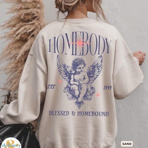May include: Sand-colored sweatshirt with the word "HOMEBODY" in purple script across the back, along with an angel graphic and the text "BLESSED & HOMEBOUND". The sweatshirt also has the text "EST 1999".