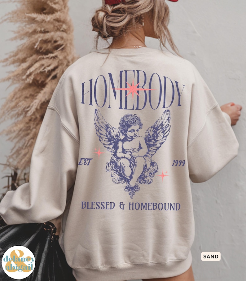 May include: Sand-colored sweatshirt with the word "HOMEBODY" in purple script across the back, along with an angel graphic and the text "BLESSED & HOMEBOUND". The sweatshirt also has the text "EST 1999".