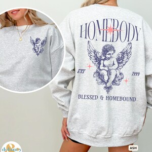 May include: A light grey sweatshirt with a cherub graphic on the front and back. The back features the text "HOMEBODY" above the cherub, with "BLESSED & HOMEBOUND" below. The sweatshirt has a relaxed fit and long sleeves.