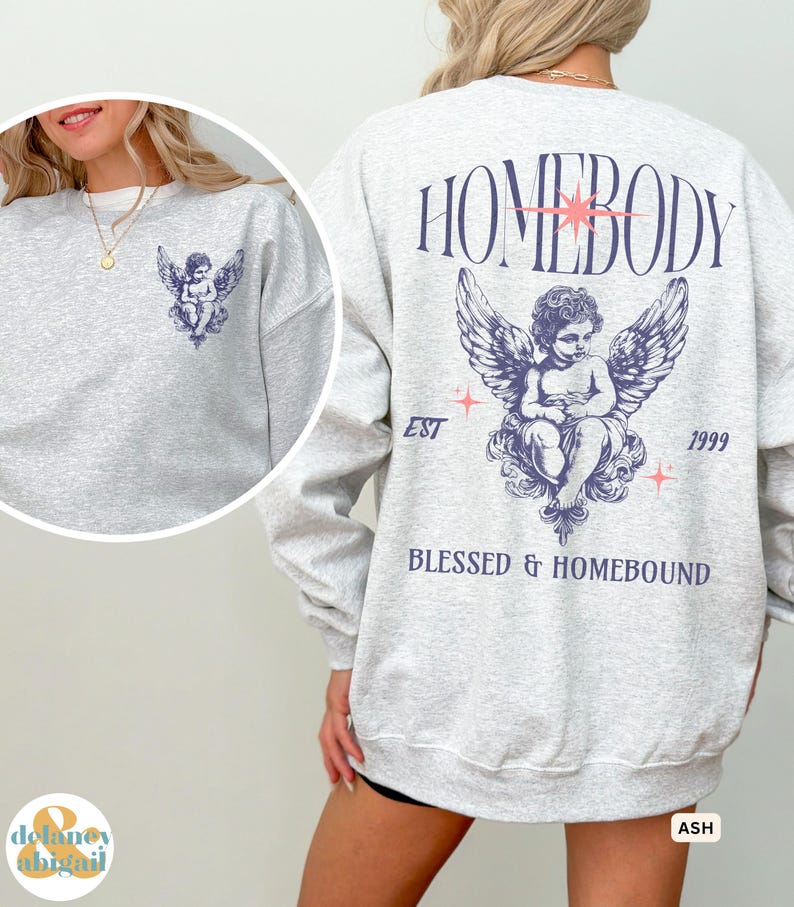 May include: A light grey sweatshirt with a cherub graphic on the front and back. The back features the text "HOMEBODY" above the cherub, with "BLESSED & HOMEBOUND" below. The sweatshirt has a relaxed fit and long sleeves.