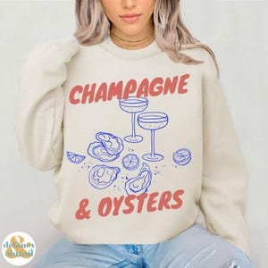 May include: A cream colored sweatshirt with the text "Champagne & Oysters" in red and blue lettering. The design includes two glasses of champagne and a cluster of oysters.