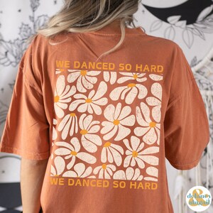 May include: A burnt orange t-shirt with a white daisy print and the text "We danced so hard" on the back.