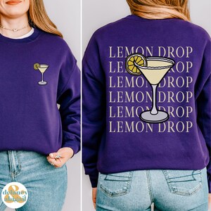 Pu&ograve; includere: Felpa viola con il testo "Lemon Drop" ripetuto cinque volte e una grafica di un bicchiere di martini al limone.