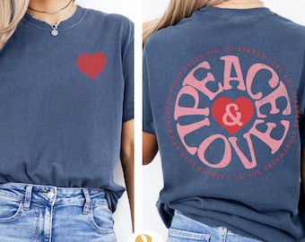 May include: A navy blue t-shirt with a pink and red peace and love graphic on the back and a red heart on the front. The shirt is being modeled by a person wearing blue jeans.