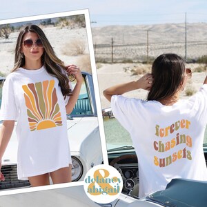 May include: White t-shirt with a retro-style sun graphic in shades of orange, yellow, and brown. The back of the shirt features the text "forever chasing sunsets" in a matching color palette. The shirt is worn outdoors.