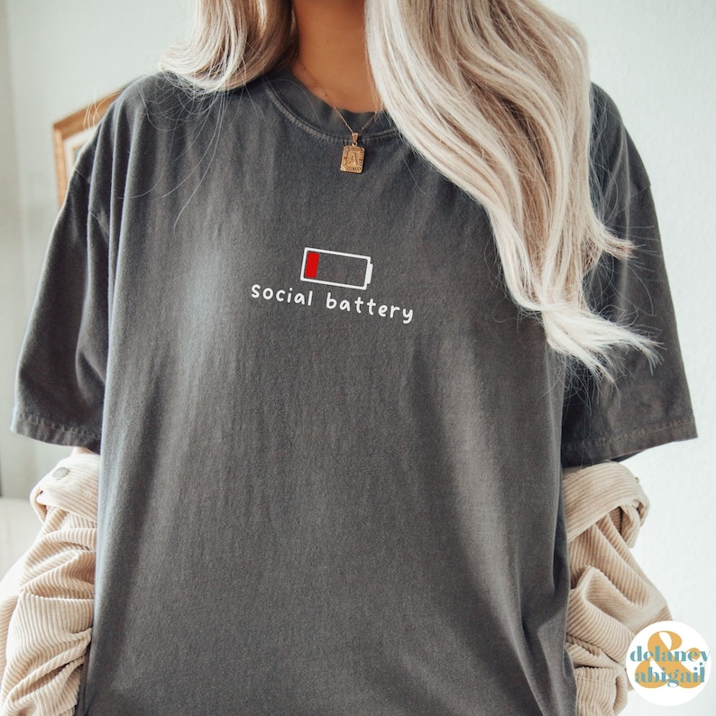 May include: A gray t-shirt with a white graphic of a low battery icon and the text "social battery".