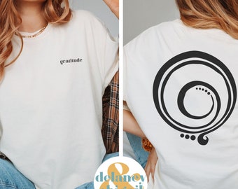 May include: Two white t-shirts with different designs. One shirt has the word "gratitude" printed on the front. The other shirt has a black and white abstract design on the back. Both shirts have the words "Delaney & Abigail" printed on the front.