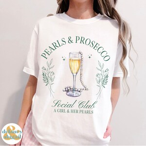 May include: White t-shirt with a graphic design featuring a champagne flute, pearls, and the text "Pearls & Prosecco Social Club: A Girl & Her Pearls" in green. The design includes floral accents.