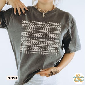 May include: Dark grey t-shirt with a white geometric design. The shirt has a crew neck and short sleeves. The word "PEPPER" is printed in white on the bottom left corner of the shirt.