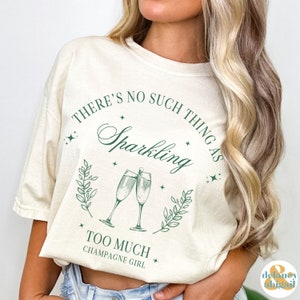 May include: A beige t-shirt with a green graphic design that says "There's no such thing as sparkling too much champagne girl". The graphic features two champagne glasses with green leaves and stars.