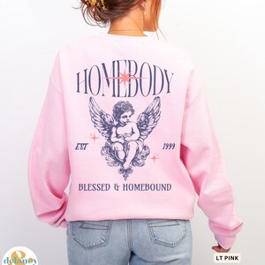May include: Light pink sweatshirt with a graphic on the back featuring the word "HOMEBODY" above an angel illustration with wings. The text "EST 1999" and "BLESSED & HOMEBOUND" are also printed on the sweatshirt.