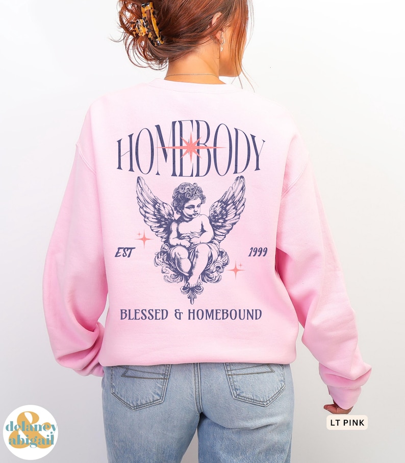 May include: Light pink sweatshirt with a graphic on the back featuring the word "HOMEBODY" above an angel illustration with wings. The text "EST 1999" and "BLESSED & HOMEBOUND" are also printed on the sweatshirt.