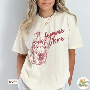May include: Ivory-colored t-shirt featuring a maroon graphic of a cowgirl riding a bear, with the words "femme libre" in maroon script. The shirt has short sleeves and a crew neck. The word "IVORY" is printed in a white box at the bottom left.
