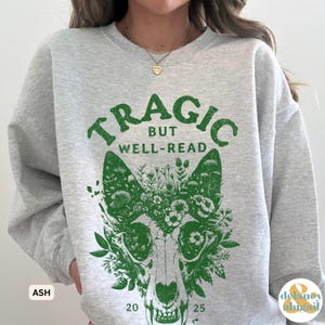 May include: Ash gray sweatshirt with the text "TRAGIC BUT WELL-READ" in green above a green floral wolf skull design. The numbers "20" and "25" are below the skull. The sweatshirt has a crew neck and long sleeves.