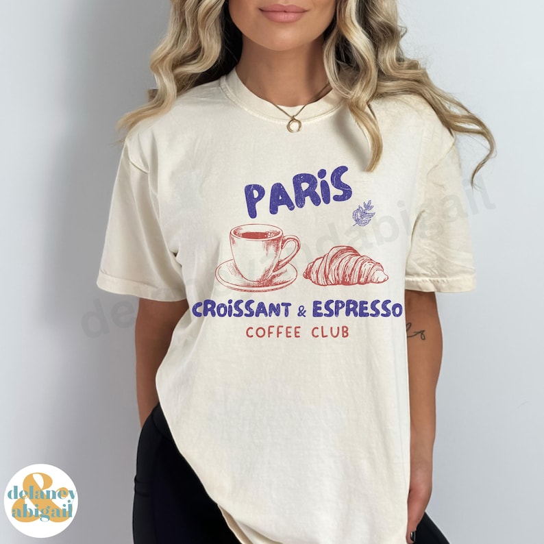 May include: A cream-colored t-shirt with a graphic design featuring a cup of coffee, a croissant, and the text "Paris Croissant & Espresso Coffee Club".