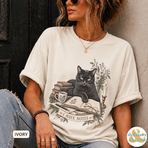 May include: An ivory t-shirt featuring a graphic of a black cat resting on a stack of books next to a mug, a candle, and an open book with glasses. The text "I HAVE NOTES" is printed on a banner below the illustration, surrounded by floral accents.