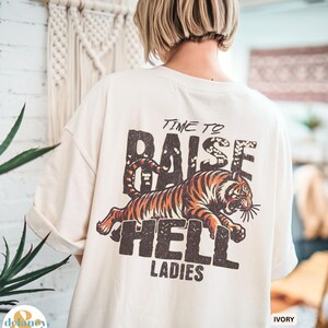 May include: Ivory-colored t-shirt with rolled-up sleeves. The back of the shirt features a distressed graphic of a tiger and the words "TIME TO RAISE HELL LADIES" in a bold, vintage font. The shirt is a relaxed fit.