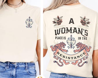 May include: A cream-colored t-shirt with a graphic design featuring two tigers, a heart with a sword, and the text "A Woman's Place is in the Resistance".