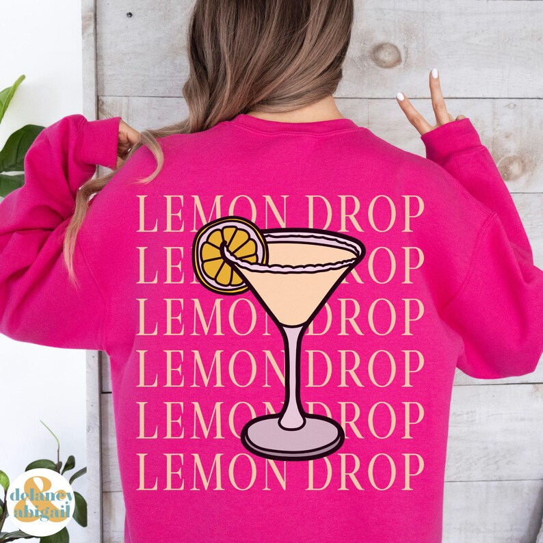 Pu&ograve; includere: Felpa rosa con il testo "Lemon Drop" ripetuto pi&ugrave; volte in un rosa chiaro. Un bicchiere da cocktail rosa con una fetta di limone giallo &egrave; centrato sulla felpa.