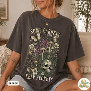 May include: A charcoal grey t-shirt featuring a graphic print with the text SOME GARDENS KEEP SECRETS. The design includes a human skull, mushrooms, a coiled snake, butterflies, and purple foxglove flowers in shades of green, white, and purple.