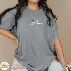 May include: A gray t-shirt with a white graphic of a low battery icon and the text "social battery".