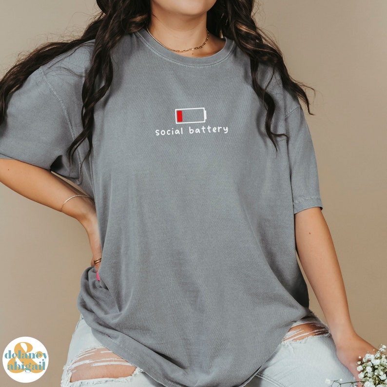 May include: A gray t-shirt with a white graphic of a low battery icon and the text "social battery".