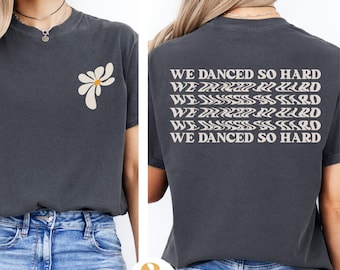 May include: A dark gray t-shirt with a white daisy on the front and the text "WE DANCED SO HARD" repeated multiple times on the back in a wavy font.