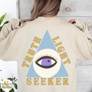 May include: A beige sweatshirt with a light blue triangle and a stylized eye inside. The triangle has the words "Truth" and "Light" on either side and the word "Seeker" at the bottom.