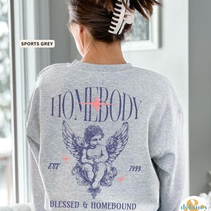 May include: A light gray sweatshirt with the word "HOMEBODY" in a stylized font, a cherub with wings, and the text "BLESSED & HOMEBOUND". The sweatshirt also has the text "EST 1999" and the words "SPORTS GREY".