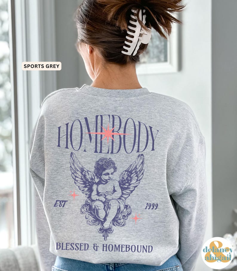 May include: A light gray sweatshirt with the word "HOMEBODY" in a stylized font, a cherub with wings, and the text "BLESSED & HOMEBOUND". The sweatshirt also has the text "EST 1999" and the words "SPORTS GREY".