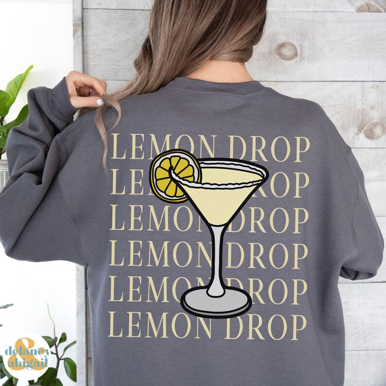 Pu&ograve; includere: Felpa grigia con il testo "Lemon Drop" ripetuto cinque volte in una colonna verticale. Una fetta di limone gialla e un bicchiere da martini con un liquido giallo sono centrati sulla felpa.