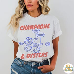 May include: White t-shirt with blue text that reads "Champagne & Oysters". The text is in a fun, casual font. There are blue illustrations of champagne flutes and oysters on the shirt.