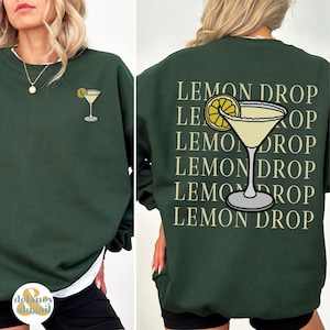 Pu&ograve; includere: Una felpa verde scuro con il testo "Lemon Drop" ripetuto cinque volte sul retro. Una fetta di limone giallo e un bicchiere da martini con un liquido giallo sono stampati sul retro della felpa. Un bicchiere da martini pi&ugrave; piccolo con un liquido giallo &egrave; stampato sul davanti della felpa.