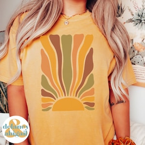 May include: Mustard yellow t-shirt featuring a retro-style sunburst design in shades of orange, brown, and green. The graphic is centered on the front of the shirt. The shirt has short sleeves and a crew neck.