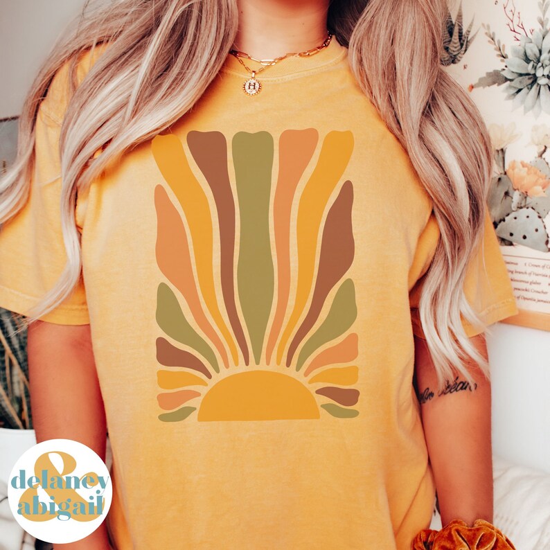 May include: Mustard yellow t-shirt featuring a retro-style sunburst design in shades of orange, brown, and green. The graphic is centered on the front of the shirt. The shirt has short sleeves and a crew neck.