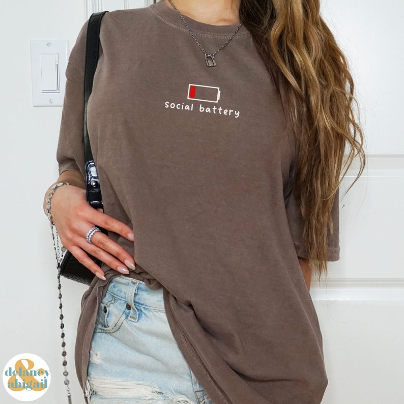 May include: A brown oversized t-shirt with a graphic of a low battery icon and the text "social battery".