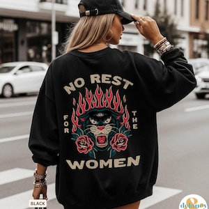 May include: Black sweatshirt with a graphic design on the back. The design features a panther with flames, roses, and the text "NO REST FOR THE WOMEN". The sweatshirt is worn by a person wearing a black cap.