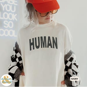 May include: Ivory-colored long-sleeve shirt with the word "HUMAN" printed in black block letters. The person is wearing an orange baseball cap and sunglasses. A black and white checkered sweater is draped over the shoulders.