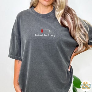 May include: A dark gray oversized t-shirt with a white graphic of a low battery icon and the text "social battery".