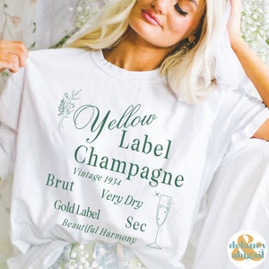 May include: White t-shirt with green text that reads "Yellow Label Champagne Vintage 1934 Brut Very Dry Sec Gold Label Beautiful Harmony". A green line drawing of a champagne flute is to the right of the text.